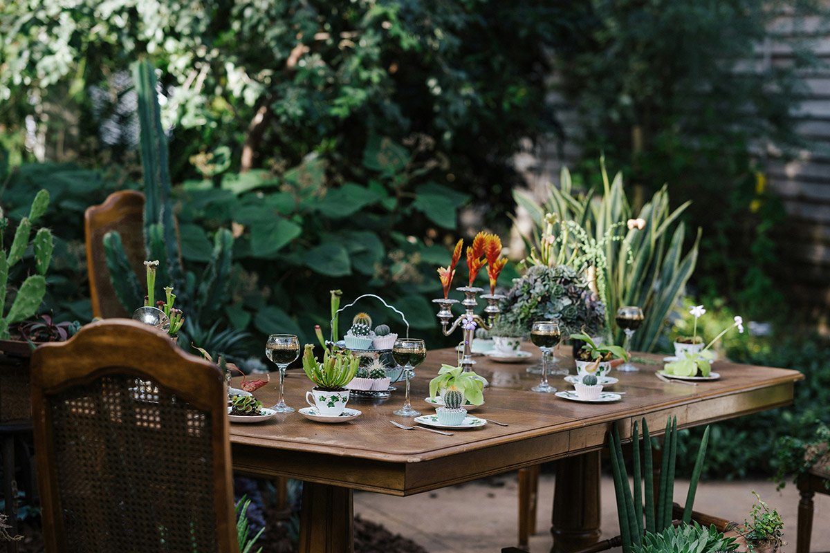 Mesa de madera al aire libre decorada con copas, candelabros y plantas suculentas, rodeada de vegetación exuberante en un jardín